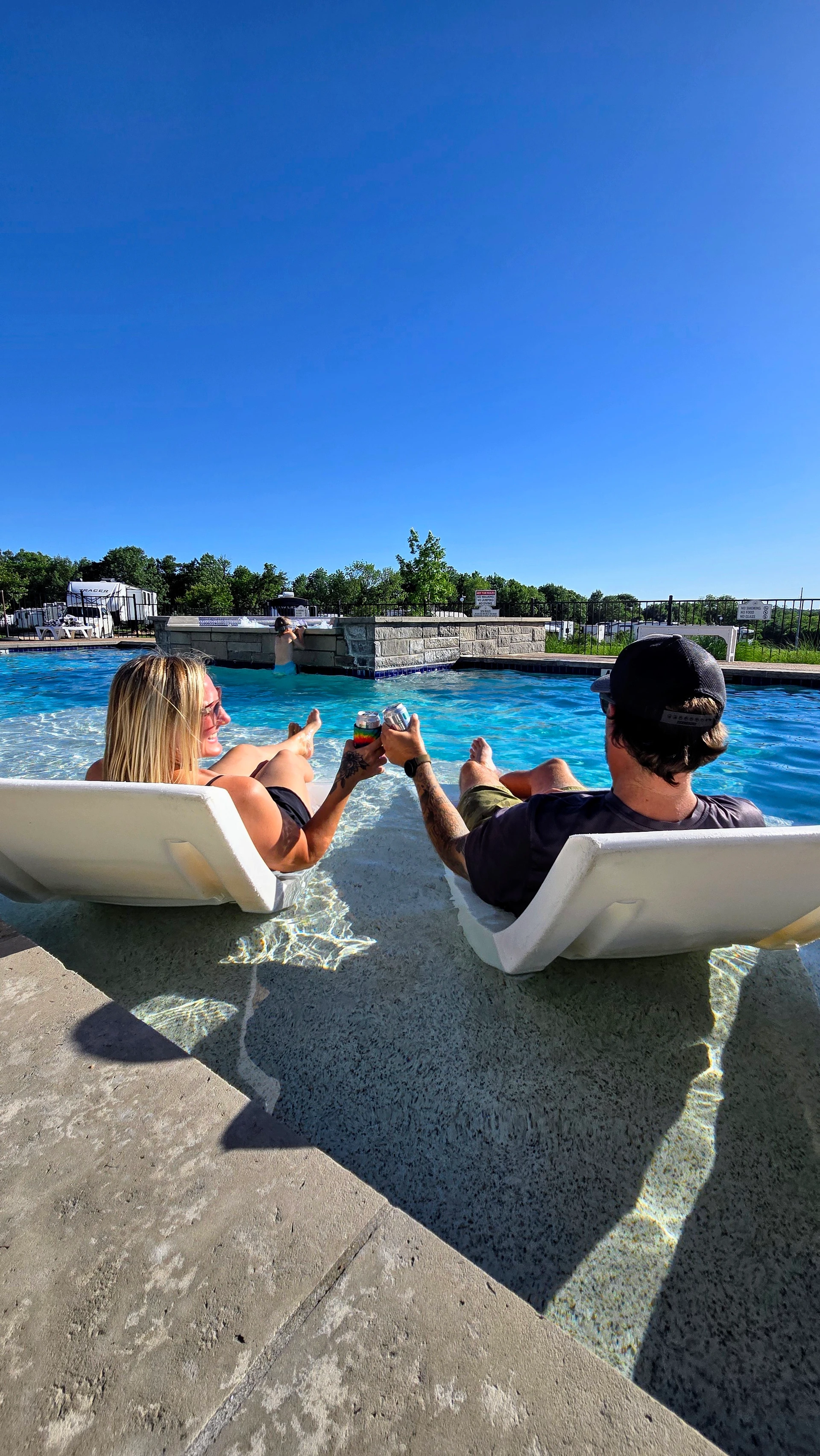 Guests relaxing in in-pool lounge chairs on the sun shelf, toasting drinks in clear blue water at Sugar Hill RV Resort.