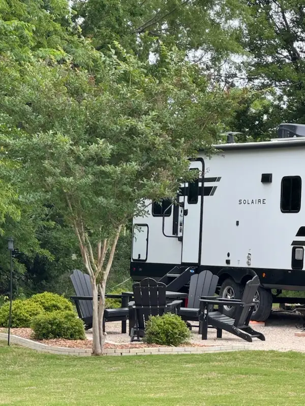 Landscaped RV site with brick-edged patio, black Adirondack chairs around a table, and a travel trailer backed to a green treeline at Sugar Hill RV Resort.